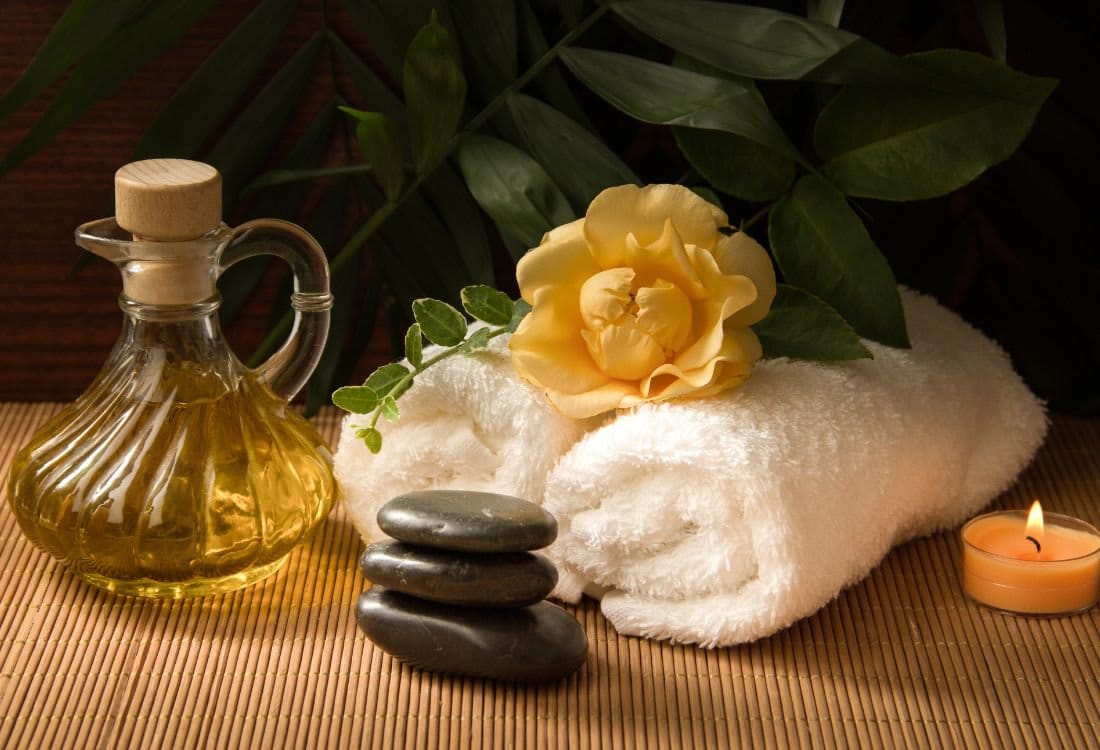 Spa products including massage oil bottle, white towel, black hot stones, yellow rose, and lit candle on bamboo mat.