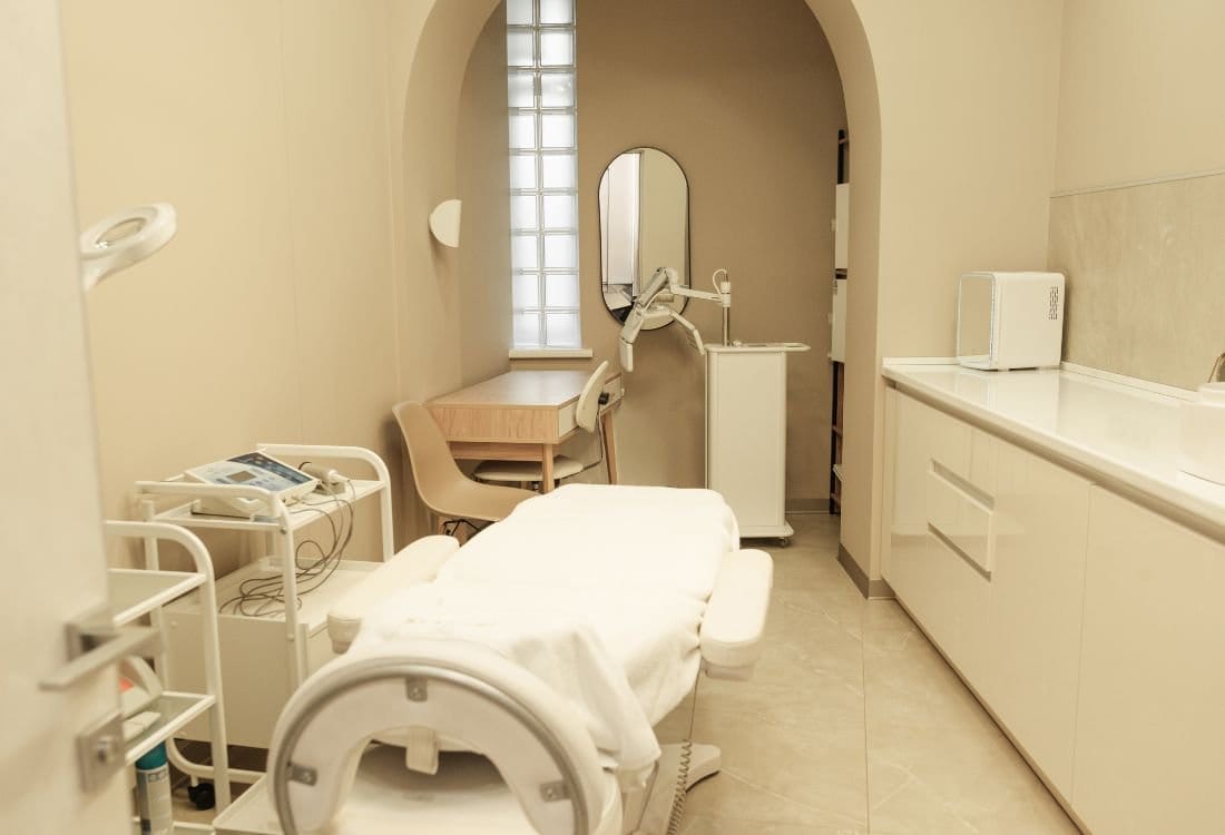 Spa treatment room with massage table, arched window, mirror, and cream-colored cabinetry. Spa treatment room with massage table, arched window, mirror, and cream-colored cabinetry.